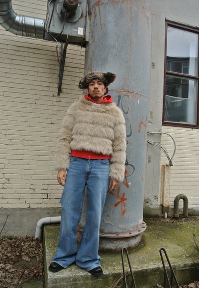 Chaqueta de piel sintética con tiras horizontales en beige claro, superpuesta a una sudadera roja, combinada con jeans de pierna ancha azules y zapatos negros.