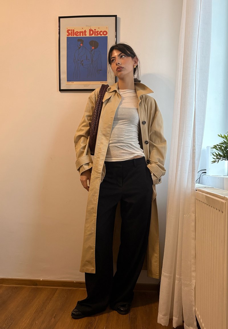 Trench beige, pantaloni neri a gamba larga e top bianco aderente; tessuto robusto, bottoni come dettagli e borsa intrecciata. Sfondo semplice con poster.