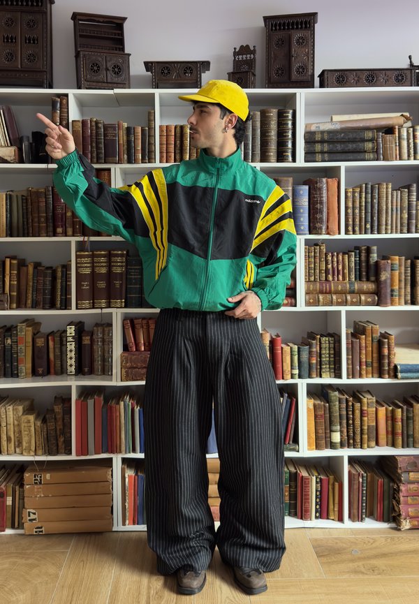 Bărbat cu șapcă galbenă, geacă verde Adidas și pantaloni negri cu dungi subțiri, care arată spre stânga, stând în fața unor rafturi pline cu cărți vechi și sculpturi din lemn.