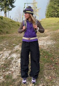 A szabadtéri ruházat egy lila polar-fleecz mellényt mutat be, amely egy barna hosszúujjú póló felett van, fekete cargo nadrággal és lila sportcipővel párosítva.