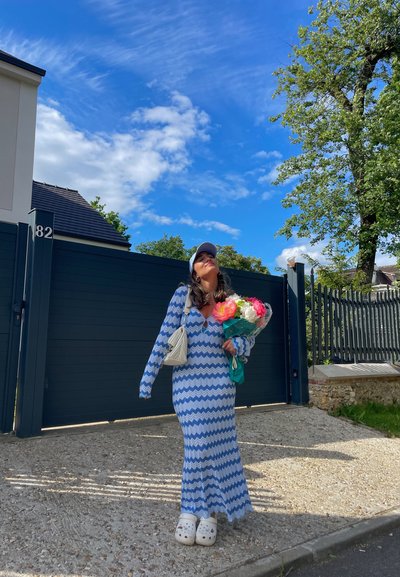 Vestido maxi de patrón zigzag en azul y blanco con mangas largas, combinado con zuecos blancos y un pequeño bolso claro, sosteniendo un ramo de flores.