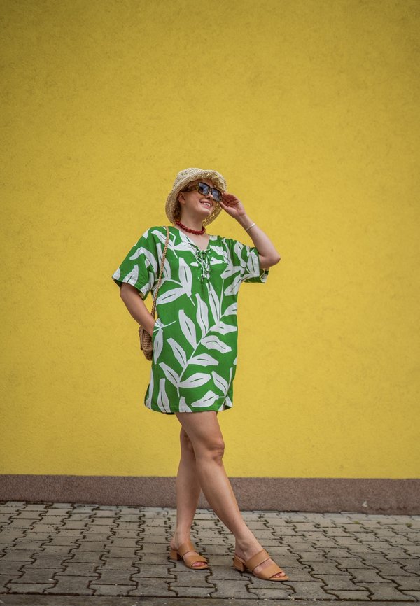 Grøn kort kjole med hvide bladmønstre, løs pasform, V-hals, accessoriseret med en stråhat, solbriller og en rød halskæde.