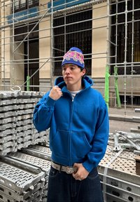 Jeune personne portant un sweat à capuche bleu et un bonnet coloré se tient près d'un échafaudage métallique empilé sur le trottoir en ville.