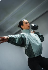 En kvinna i en grå vindjacka med vita detaljer lyfter en svart hantel. Vindjackan �är något oversize med veckade muddar och hög krage.