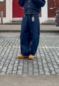 Persona con pantalones anchos azules y botas color crema de pie en una calle de adoquines frente a puertas rojas de un edificio.