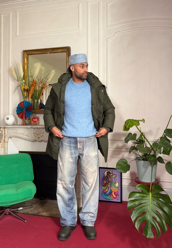 Homme en pull bleu, veste verte et jean délavé se tient dans une pièce avec moquette rouge, fauteuil vert, grande plante et œuvre d'art encadrée colorée.