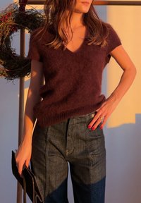 Femme portant un pull marron à manches courtes et duveteux et un jean foncé taille haute, tenant une pochette noire, avec une couronne en arrière-plan.