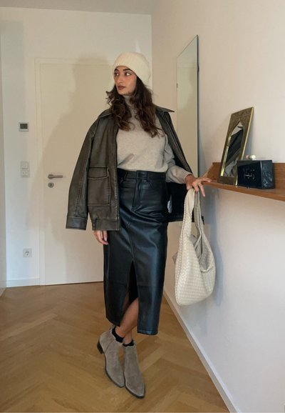 Chaqueta de cuero caqui, suéter gris, falda lápiz de cuero negro con abertura frontal, botines grises y bolso de mano de mimbre beige. Suelo de madera.