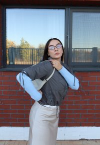 Femeie cu ochelari, purtând o jachetă gri și o fustă bej, ajustând bretelele jachetei în fața unui perete de cărămidă roșie și a unei ferestre.