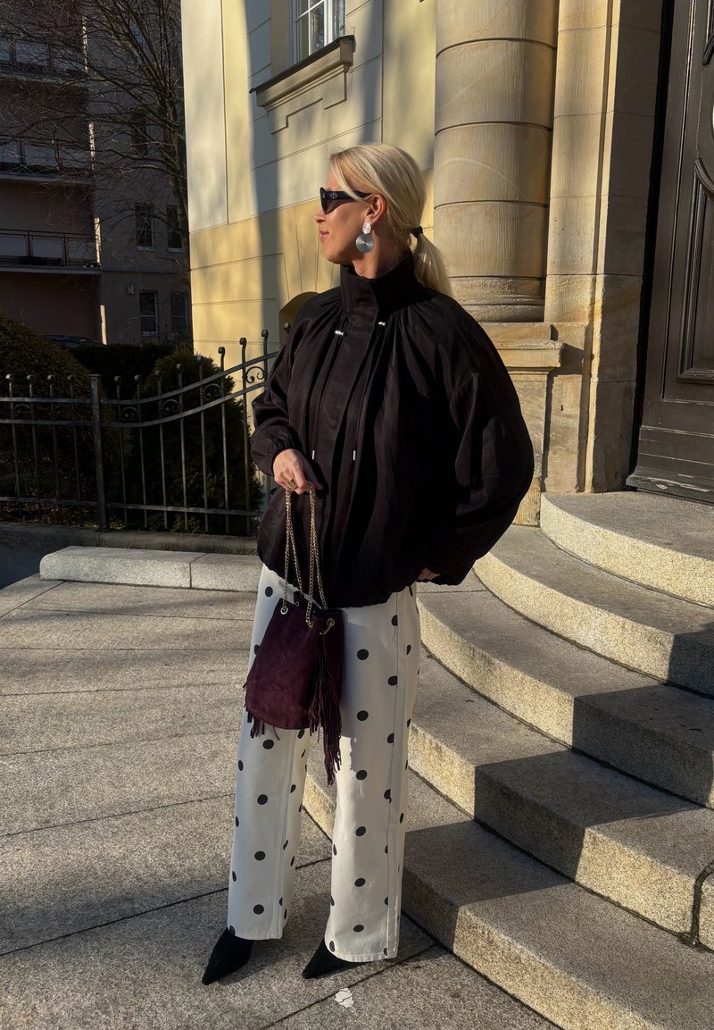 Femme aux cheveux blonds portant une veste noire, un pantalon blanc à pois, des chaussures noires et des lunettes de soleil, tenant un sac violet, debout sur des marches en pierre en plein air.