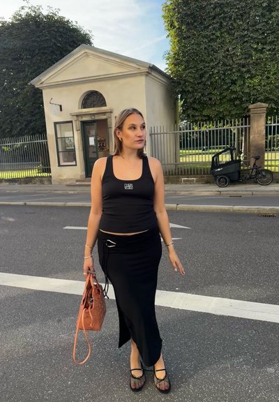 Top negro sin mangas con un logo, falda negra ajustada con una abertura lateral, zapatos de malla negros y un bolso de mano tejido naranja. Escenario urbano.