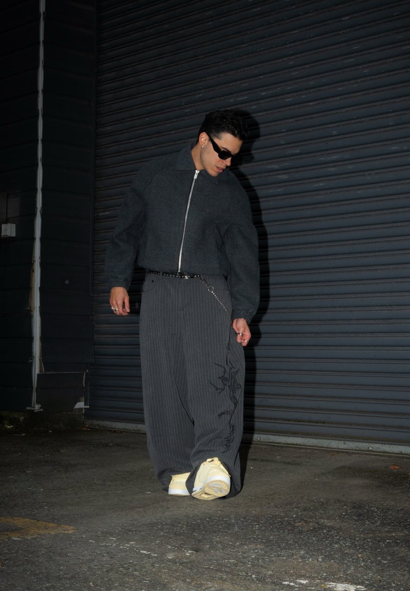 Dark grey cropped jacket with a zip front, wide grey-striped trousers featuring a graphic, and light-coloured sneakers on a textured surface.