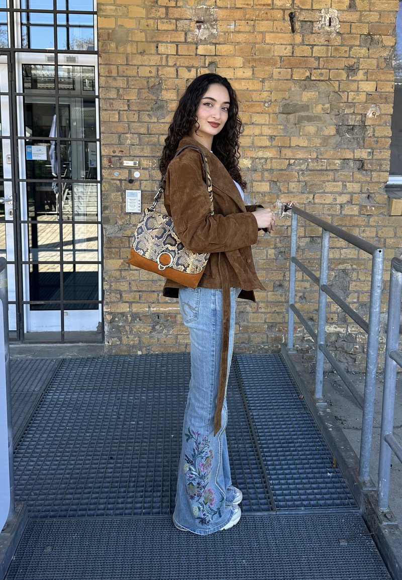 Giovane donna con capelli ricci che indossa una giacca marrone, jeans ricamati, sneakers bianchi e una borsa con motivo a serpente davanti a un muro di mattoni.