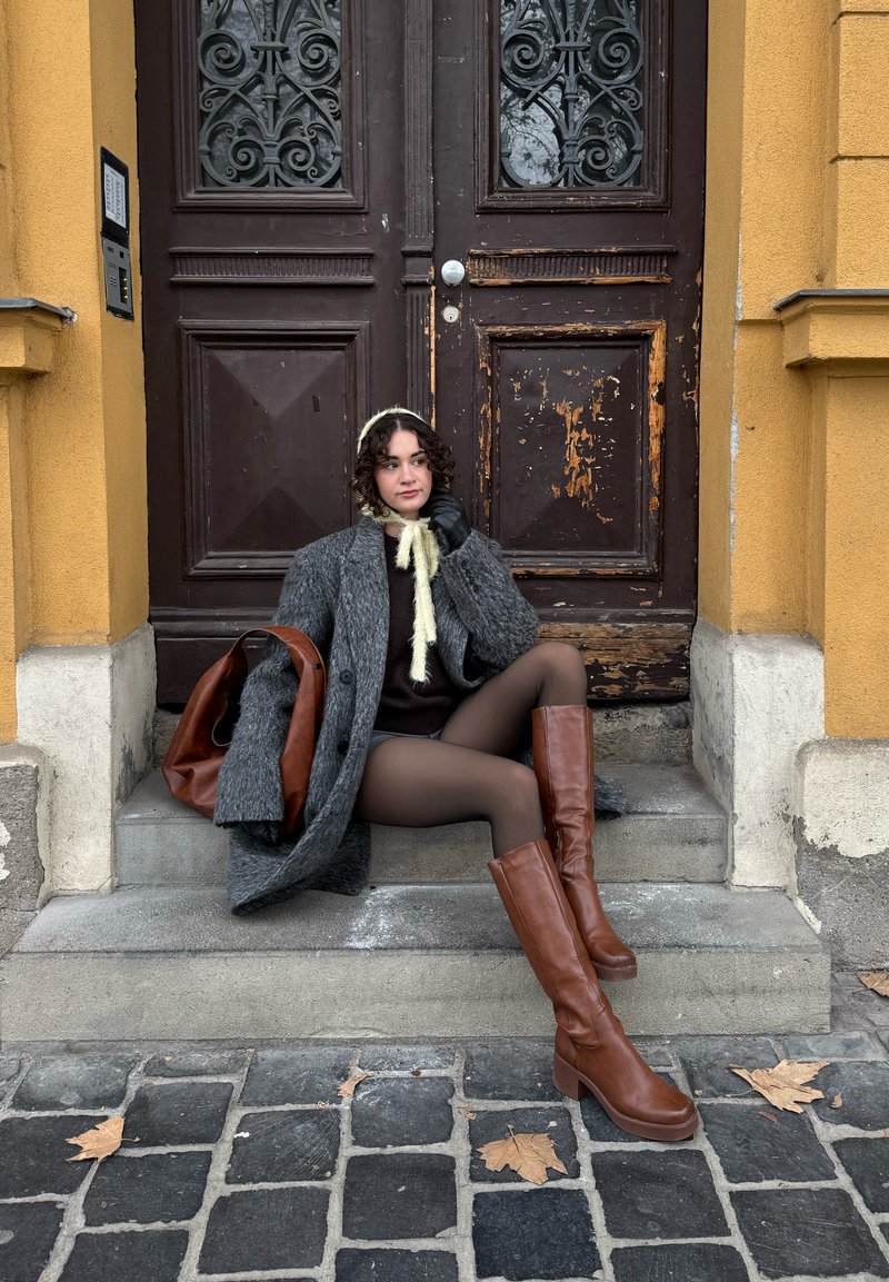 Donna con stivali marroni, collant, cappotto grigio e cuffia color crema seduta sui gradini di pietra davanti a porte doppie di legno scuro consumato su edificio giallo.