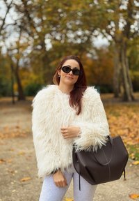 Biela umelá kožušinová bunda s nadýchanou textúrou, sivé legíny a veľká tmavohnedá kabelka. Hladký, štruktúrovaný dizajn s viditeľnými švovými akcentmi.