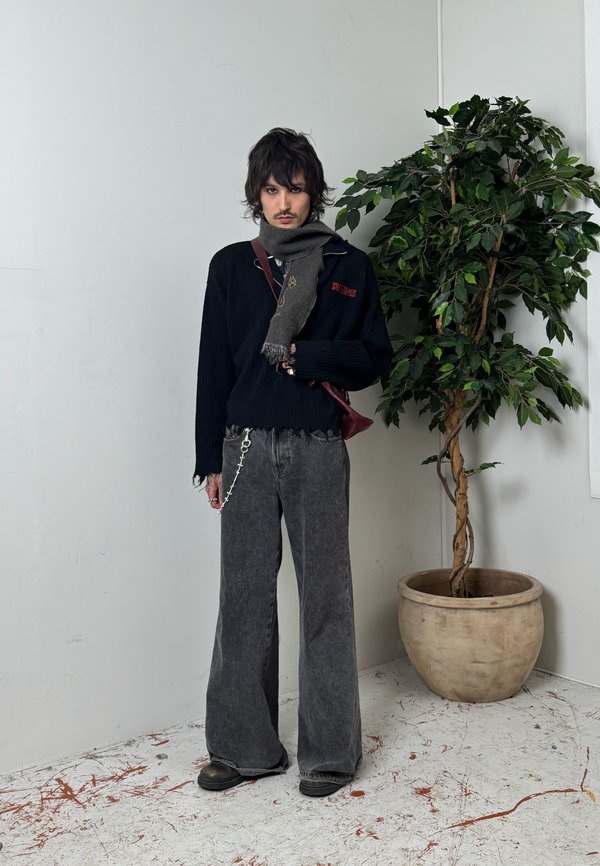 Noor inimene, kes kannab musta kampsunit, halli laia säärega teksaseid, halli salli ja musti kingi, seisab suure toalille pottide kõrval.