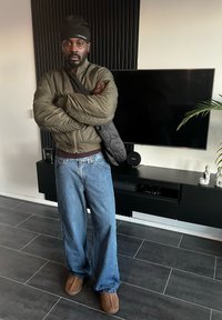 Veste matelassée vert olive, jean bleu clair ample, chaussures marron et bonnet noir. Debout à l'intérieur, les bras croisés, avec une décoration moderne.