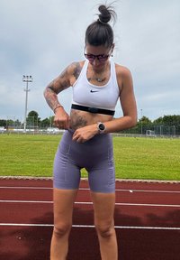 White sports bra with black logo, purple high-waisted shorts, and a fitness tracker; outdoors on a track with grass in the background.