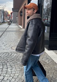 Casaco de pele com colarinho de pelúcia, cor castanha, usado sobre uma camisa branca e jeans azul claro, numa rua de calçada.