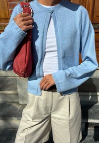 Cardigan azul claro feito de um fio macio, com decote redondo e fecho de botão, combinado com uma blusa branca e calções bege. Uma bolsa vermelha é segurada.