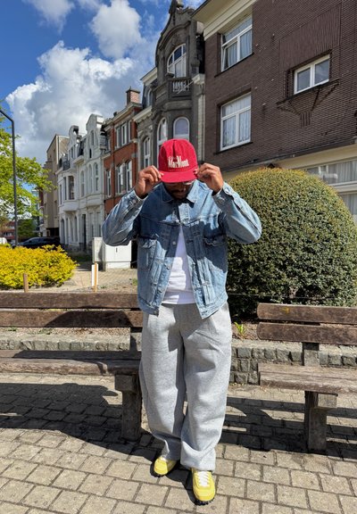 Hombre con gorra roja, chaqueta vaquera, pantalones de chándal grises y zapatillas amarillas de pie junto a bancos de madera en una calle residencial soleada.