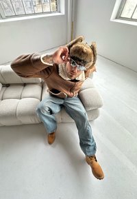 Giacca di pelle marrone con fodera in shearling, cappello di pelliccia, occhiali da sole, jeans blu chiaro e stivali color beige, seduto su un soffice divano di colore chiaro.
