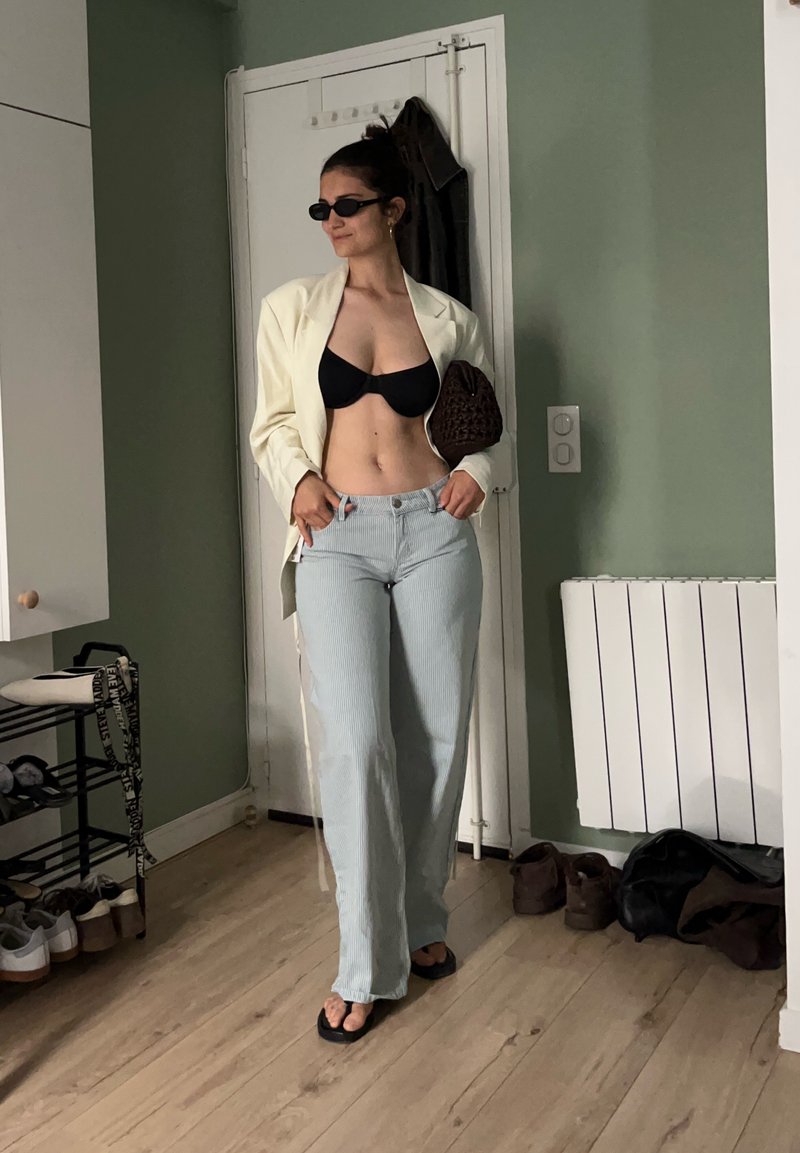 Femme portant un haut de bikini noir, un blazer crème, un pantalon rayé, des tongs et des lunettes de soleil, debout dans une pièce avec un sol en bois et des murs verts.