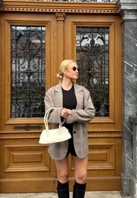 Vrouw met blond haar in een bun, die een zonnebril, een geruite blazer, een zwarte jurk draagt en een witte handtas vasthoudt, staande voor versierde houten dubbele deuren.
