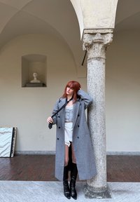 Cappotto di lana grigio con bottoni neri, indossato sopra un top grigio chiaro e una gonna bianca. Stivali neri fino al ginocchio. Colonna in pietra sullo sfondo.