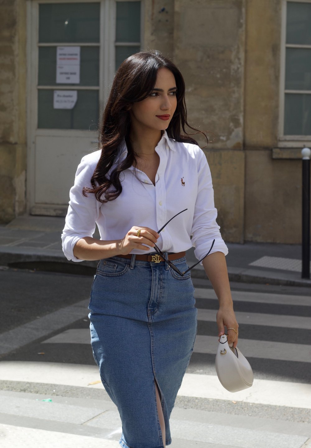 Witte blouse met knopen, spijkerrok met een voorkapesluiting, bruine riem, zwart zonnebril vasthoudend en een kleine witte handtas, wandelend op een straat.