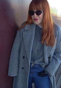 Kobieta z długimi rudymi włosami, nosząca czarne okulary przeciwsłoneczne, szary wełniany płaszcz, szary kardigan, niebieskie jeansy i czarną rękawiczkę, stoi oparta o ścianę.