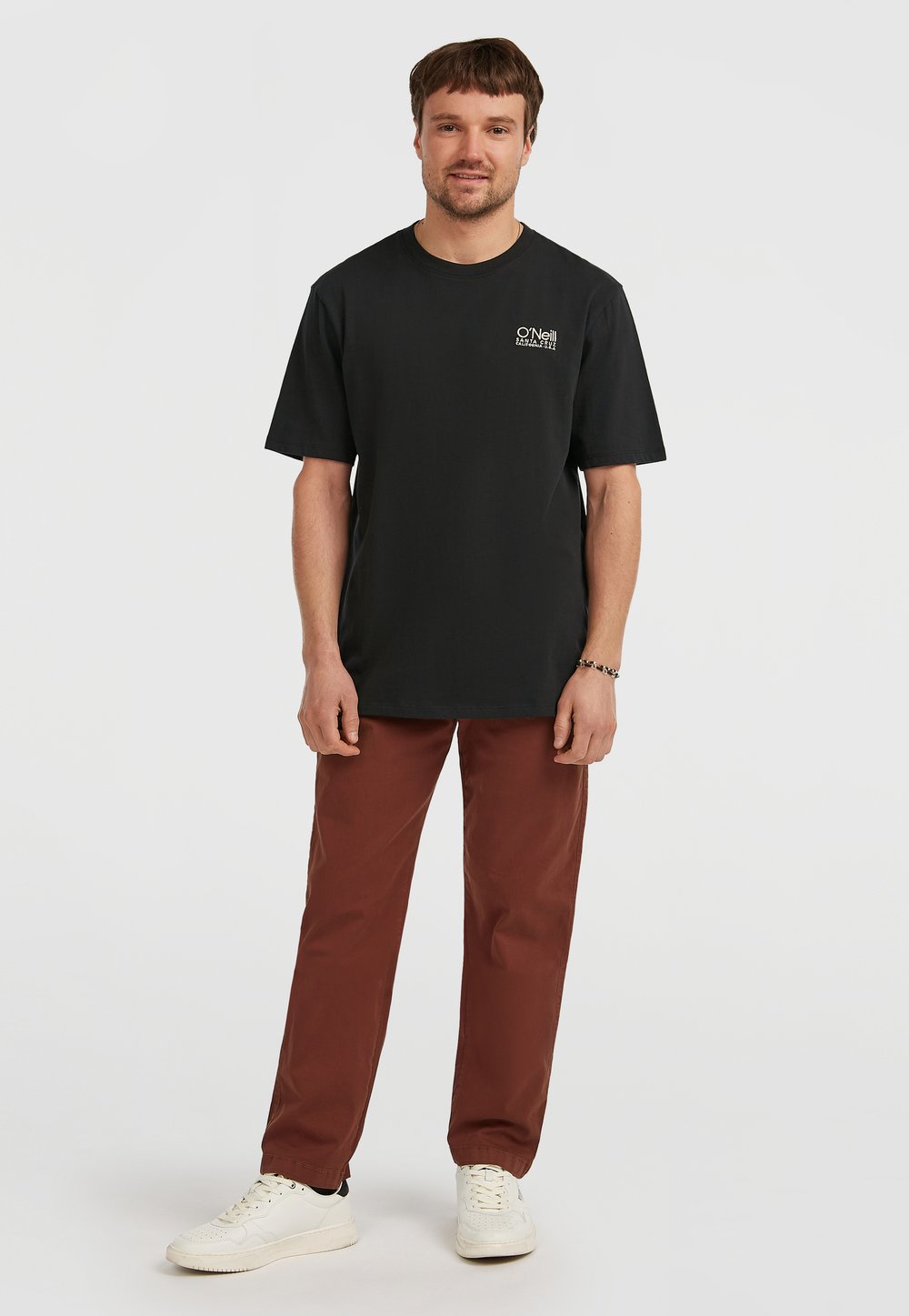 Černé tričko s krátkým rukávem s malým bílým logem na levé straně hrudi, doplněné hnědými kalhotami a bílými teniskami.