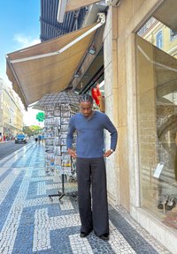 Karališkai mėlynas raštuotas megztinis, laisvas kirpimas; juodos laisvos kelnės; glotnus paviršius; stovi ant raštuoto akmens šaligatvio šalia skėčio laikiklio.