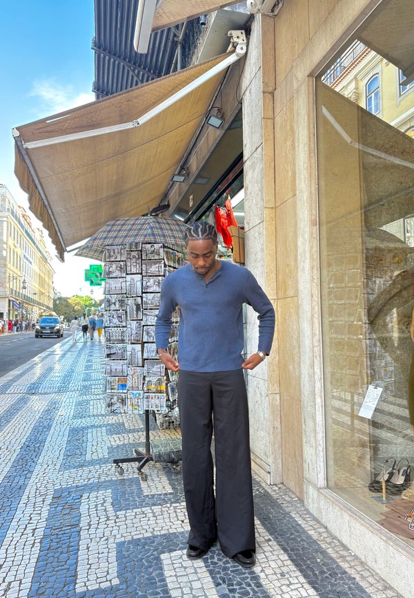Navy ribbed sweater, relaxed fit; black loose trousers; smooth texture; standing on patterned stone pavement next to an umbrella stand.