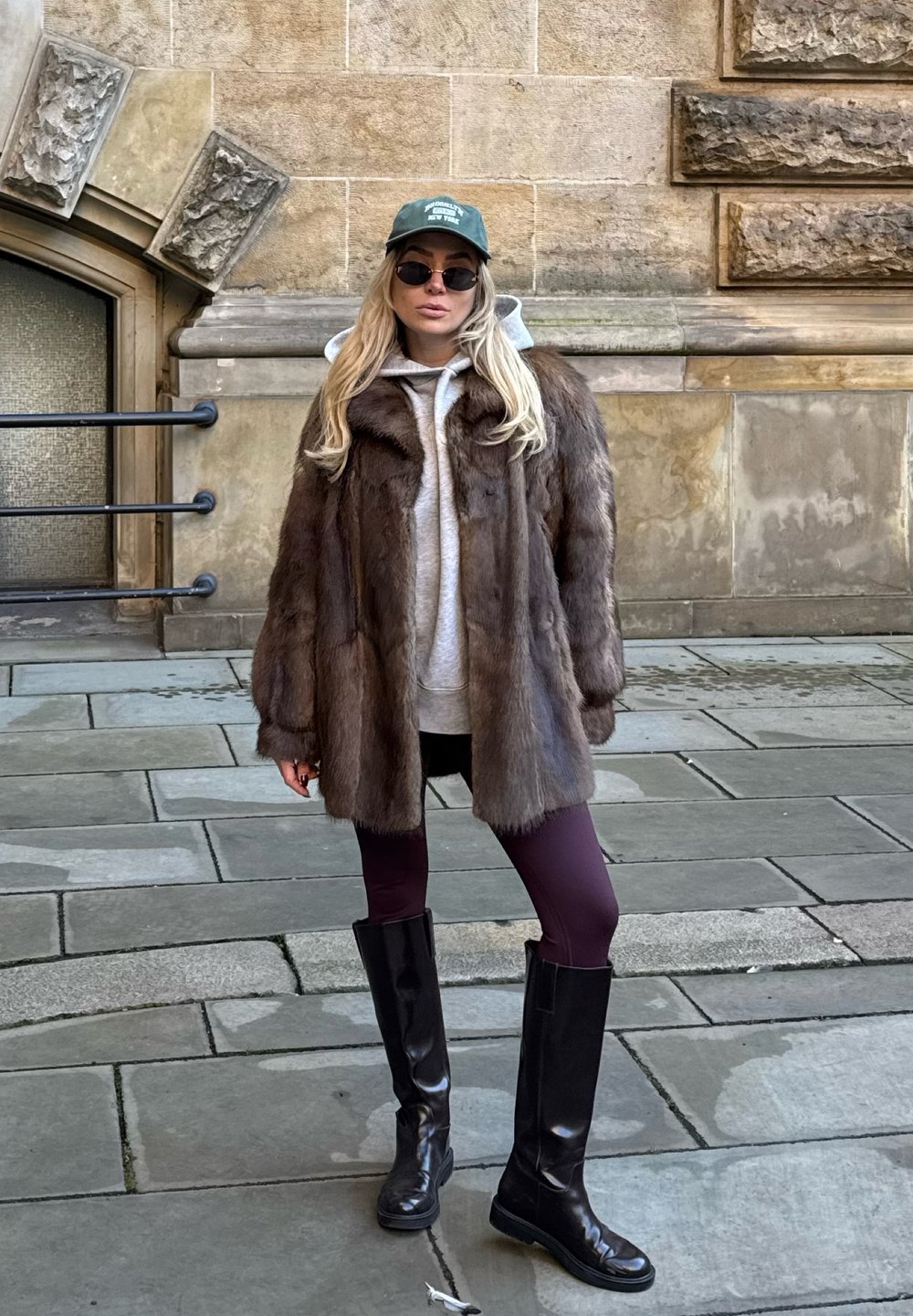 Manteau en fourrure marron sur un sweat à capuche gris, associé à des leggings bordeaux et de grandes bottes noires. Accessoires : une casquette verte et des lunettes de soleil.