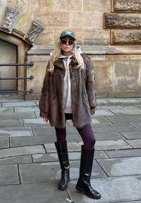 Manteau en fourrure marron sur un sweat à capuche gris, associé à des leggings bordeaux et de grandes bottes noires. Accessoires : une casquette verte et des lunettes de soleil.