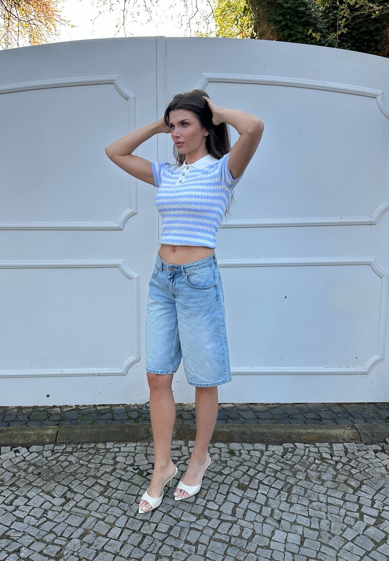 Femme ajustant ses cheveux portant un crop top rayé bleu et blanc, un short en jean jusqu'aux genoux, et des sandales blanches à talons en plein air.