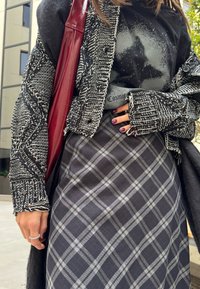 Γυναίκα που φοράει μαύρο και άσπρο πλεκτό ζακέτα, μαύρο μπλουζάκι με τύπωμα, γκρι καρό φούστα, κρατώντας κόκκινη δερμάτινη τσάντα σε εξωτερικό χώρο.