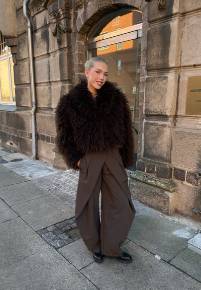 Persona de pie en una acera, vistiendo una chaqueta esponjosa marrón oscuro y pantalones marrones de pierna ancha, frente a la entrada de un edificio de piedra.