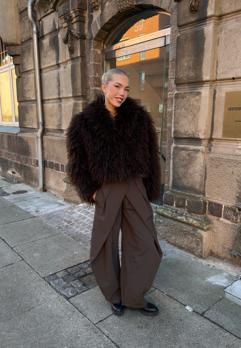 Personne debout sur un trottoir portant une veste duveteuse brun foncé et un pantalon marron à jambes larges devant l'entrée d'un bâtiment en pierre.