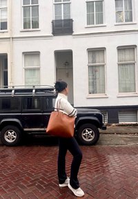 Hnědá kožená taška přes rameno s dvěma uchy, hladkým povrchem a zaobleným tvarem. Černé vozidlo s robustním designem je zaparkováno na cihlové ulici.