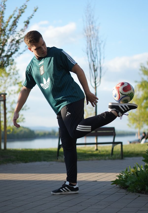 Jeune homme en tenue de sport jonglant avec un ballon de football avec son pied sur un chemin pavé près d'une rivière et des bancs de parc sous un ciel bleu.