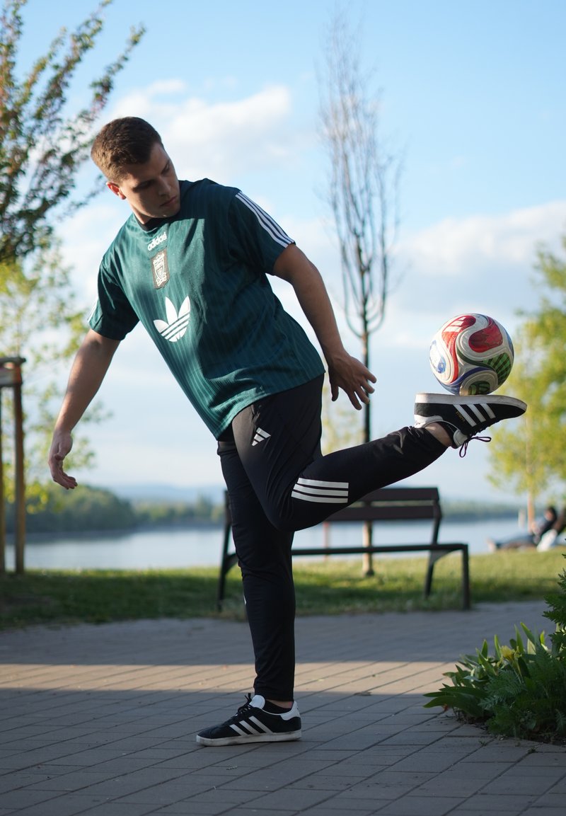 Jeune homme en tenue de sport jonglant avec un ballon de football avec son pied sur un chemin pavé près d'une rivière et des bancs de parc sous un ciel bleu.