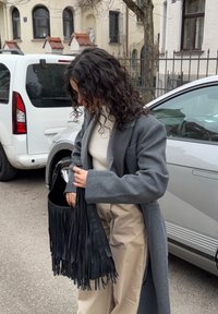 Femme aux cheveux bouclés portant un manteau gris et un pantalon beige, regardant dans un sac à main noir à franges, debout près de voitures garées dans une rue.