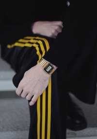 Montre numérique en or au poignet, portée avec un pantalon noir muni de bandes latérales jaunes. La montre a un cadran rectangulaire et un bracelet métallique.