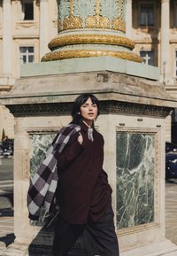 Jeune femme aux cheveux foncés se tient à côté d'un pilier en pierre orné, portant une chemise sombre, un pantalon rayé, et tenant une veste à carreaux sur l'épaule dans un cadre urbain ensoleillé.
