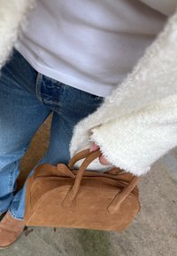 Personne portant une veste blanche en tissu duveteux, une chemise blanche et un jean bleu, tenant un sac à main en suède marron avec deux anses, debout sur un sol en béton.