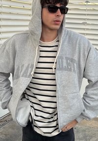 Sweatshirt cinza claro com fecho e drawstrings e bolsos frontais, usado sobre uma camisa riscada branca e preta, contra um fundo neutro.