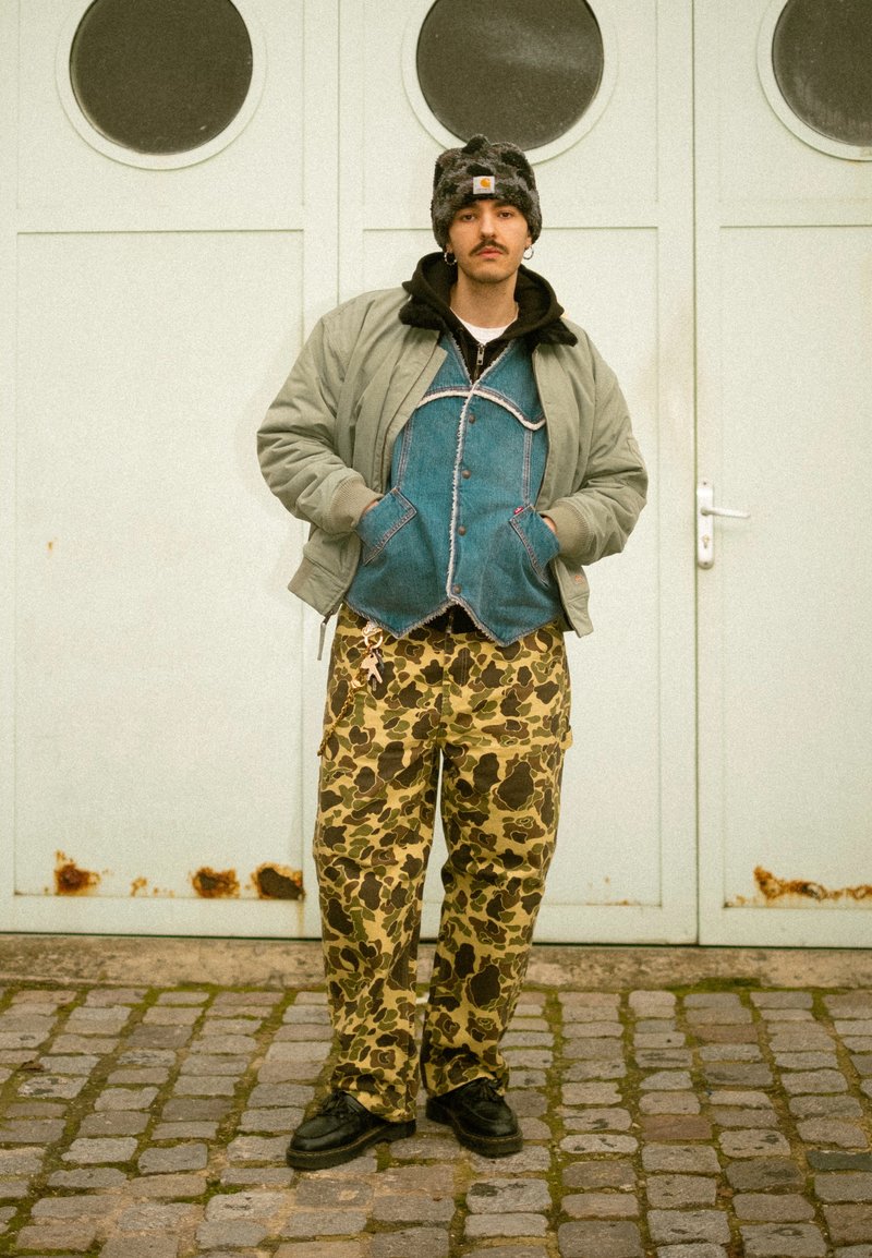 Homme avec moustache portant un bonnet noir et gris, une veste bomber grise, un gilet en jean, un pantalon camouflage et des chaussures noires, debout sur des pavés devant une porte blanche.