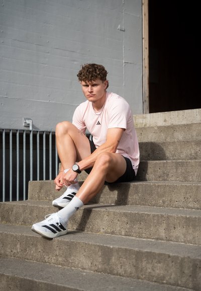 Un atleta se sienta en unos escalones de concreto, vistiendo una camiseta Adidas de color rosa claro, pantalones cortos negros, zapatillas blancas con rayas negras y calcetines blancos.